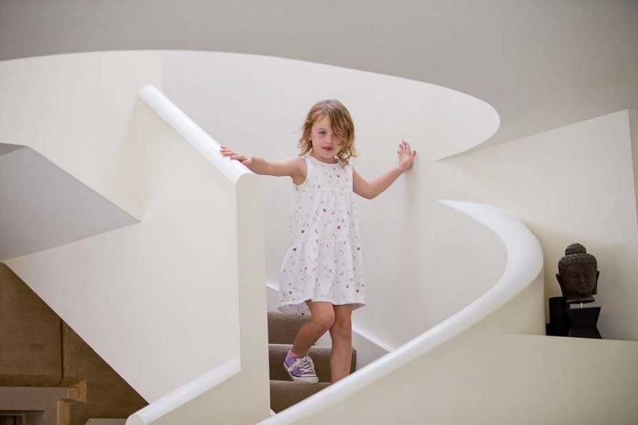 A little flower girl wearing a white dress walks down stairs at the brides home as the wedding party get ready for the wedding at Fort Takapuna and the Officers Mess in Auckland.