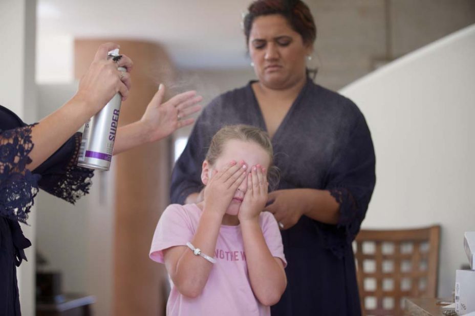 A little flower girl covers her eyes with her hands as a makeup artist sprays hairspray to fix her hairstyle as she prepares for a wedding at Fort Takapuna and the Officers Mess in Auckland.