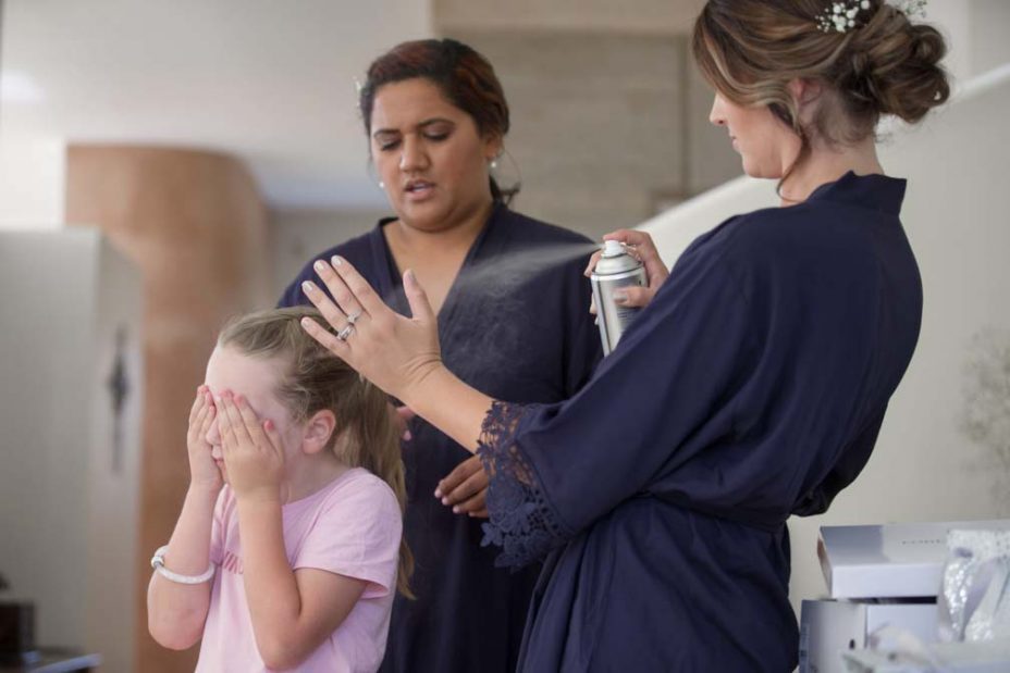 A little flower girl covers her eyes with her hands as a makeup artist sprays hairspray to fix her hairstyle as she prepares for a wedding at Fort Takapuna and the Officers Mess in Auckland.