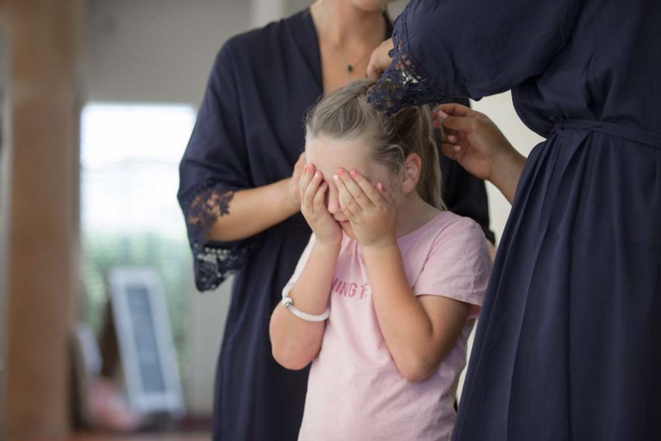 A little flower girl covers her eyes with her hands as a makeup artist sprays hairspray to fix her hairstyle as she prepares for a wedding at Fort Takapuna and the Officers Mess in Auckland.