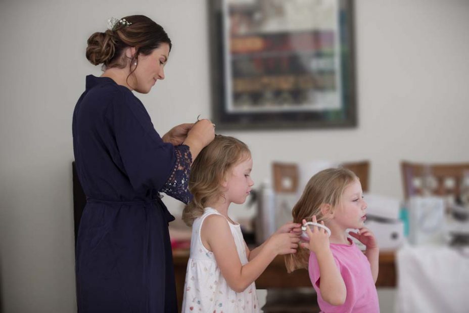 Platting their hair in a row are two little flower girls and and adult bridesmaid wearing a royal blue robe. Getting ready for the wedding at Fort Takapuna and the Officers Mess in Auckland.