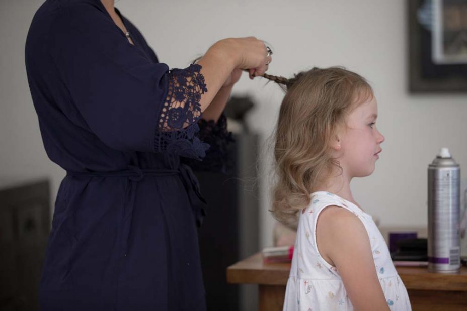 A little flower girl standing as her hair is platted by a bridesmaid wearing a royal blue robe. Getting ready for the wedding at Fort Takapuna and the Officers Mess in Auckland.