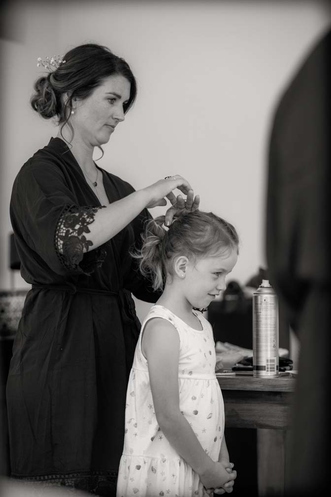 Black and white photo of a little flower girl standing as her hair is platted by a bridesmaid wearing a royal blue robe. Getting ready for the wedding at Fort Takapuna and the Officers Mess in Auckland.