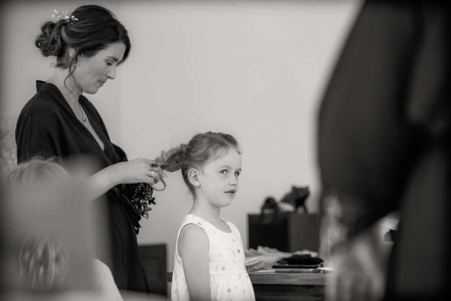 Black and white photo of a little flower girl standing as her hair is platted by a bridesmaid wearing a royal blue robe. Getting ready for the wedding at Fort Takapuna and the Officers Mess in Auckland.