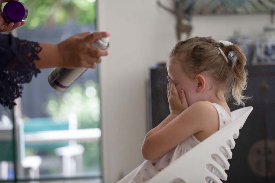 A little flower girl covers her eyes with her hands as a makeup artist sprays hairspray to fix her hairstyle as she prepares for a wedding at Fort Takapuna and the Officers Mess in Auckland.