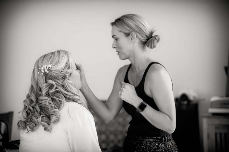 Black and white photo of a bride wearing a pink robe has her make up applied as she prepares for her wedding at Fort Takapuna and the Officers Mess in Auckland.