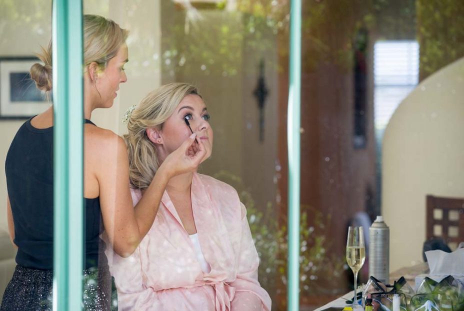 Photo through a window of a bride wearing a pink robe smiles whilst she has her make up applied whilst she prepares for her wedding at Fort Takapuna and the Officers Mess in Auckland.