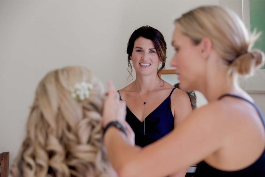 Bridesmaid wearing royal blue v-neck satin dress smiles and watches the bride wearing a pink robe have her make up applied as she prepares for her wedding at Fort Takapuna and the Officers Mess in Auckland.