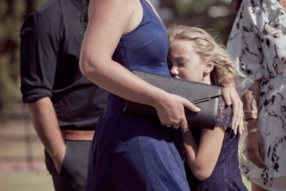 Little girl wearing a blue lace dress hugs her mother with her eyes tight shut during a wedding ceremony at Fort Takapuna in Auckland.