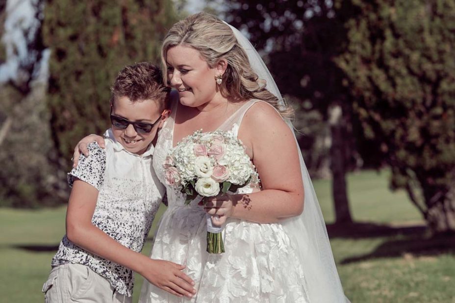 Bride wearing a full style wedding dress and holding a flower bouquet made of pink and white roses hugs a small boy wedding guest following her ceremony at Fort Takapuna in Auckland.