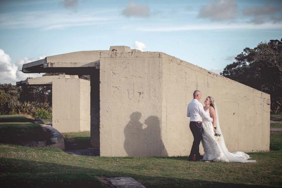 Bride wearing a full style white wedding dress holding her flower bouquet poses cuddled in with her new husband, for a creative wedding photo in front of a unique concrete structure at Fort Takapuna and the Officers Mess in Auckland