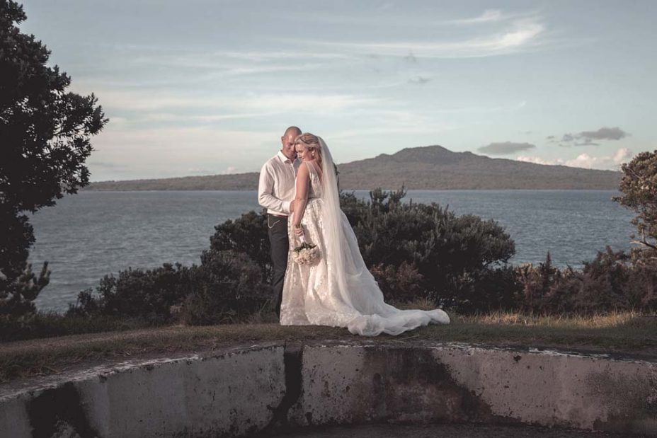 A sunny day wedding with a view over the sea to Rangitoto Island in the background. Bride wearing a full style white wedding dress with long stands with her new husband on a unique concrete structure for a creative wedding photo at Fort Takapuna and the Officers Mess in Auckland