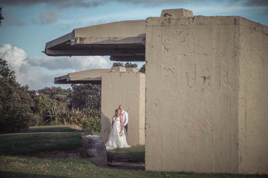 Bride wearing a full style white wedding dress holding her flower bouquet poses cuddled in with her new husband, for a creative wedding photo in front of a unique concrete structure at Fort Takapuna and the Officers Mess in Auckland