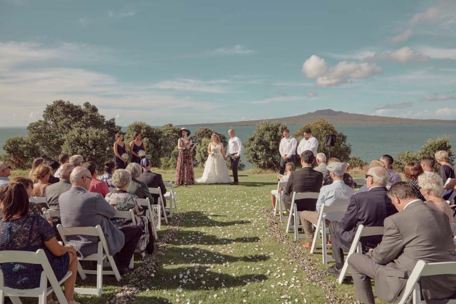 Wedding ceremony on a sunny day with a view of the sea and rangitoto Island. Guests watch as the celebrand reads to the bride wearing a full style white wedding dress and groom wearing white linen shirt and dark blue chino pants hold hand during their ceremony at Fort Takapuna and the Officers Mess in Auckland