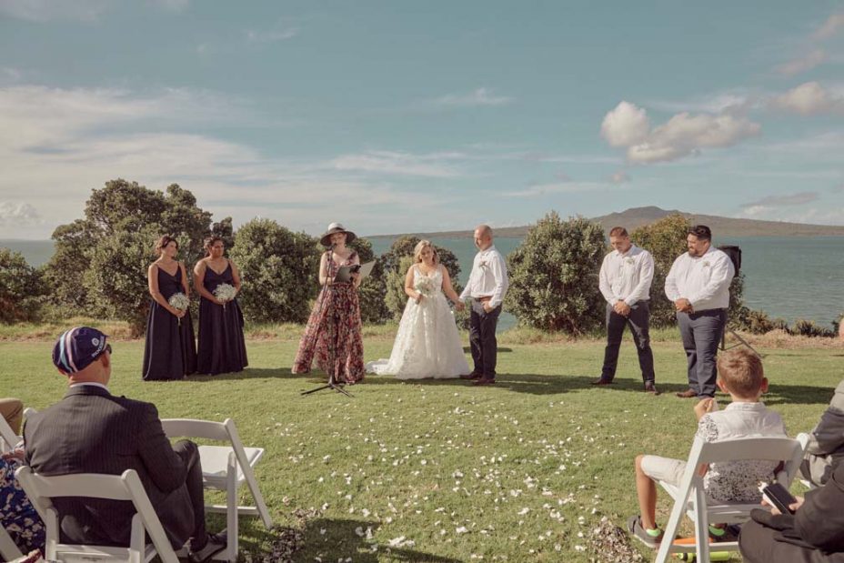 Wedding ceremony on a sunny day with a view of the sea and rangitoto Island. Guests watch as the celebrand reads to the bride wearing a full style white wedding dress and groom wearing white linen shirt and dark blue chino pants hold hand during their ceremony at Fort Takapuna and the Officers Mess in Auckland